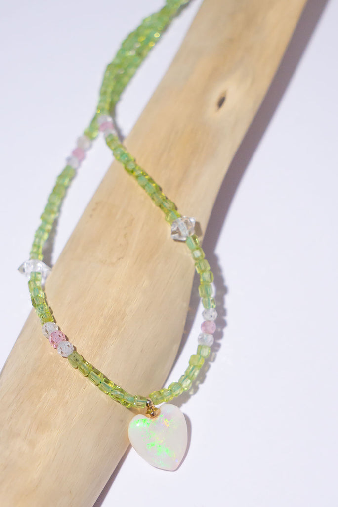 Feel the love of this beautiful crystal opal hand carved into a heart, with soft green and mauve sprinkles across the white crystal. This ethereal opal comes on a necklace of tiny faceted green Peridot beads with Herkimer Diamond and Quartz crystal highlights.